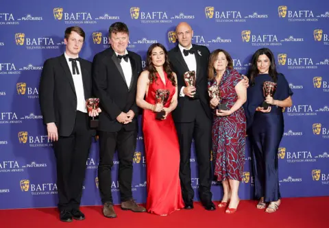 PA Media (Left to right) Ben De Pear, Ramita Navai, Karim Shah, Melanie Quigley and Menna Hijazi hold Bafta awards while looking at the camera with the Bafta logo plastered on a blue billboard behind them while on the red carpet at the event.