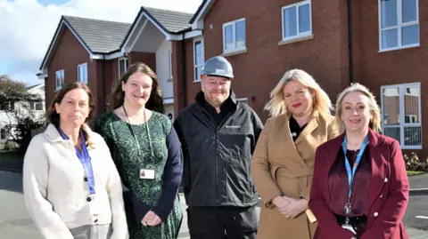 Five people standing in front of brown brick homes with pointed roofs. There are four women and one man in the middle. the two women on the left have long brown hair - one is wearing a white cardigan and the other is wearing a blue and green dress. The man is in all back and has a grey hard hat on. On the right, the two women have shoulder-length blonde hair. On the far right, the woman is wearing a maroon silk blouse and a red jacket, and the woman to the left of her is wearing a brown jacket
