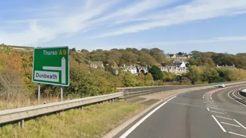 Google A green road sign on the A9 with arrows pointing north to Thurso and west to Dunbeath. A few metres along the road from the sign are houses of Dunbeath.