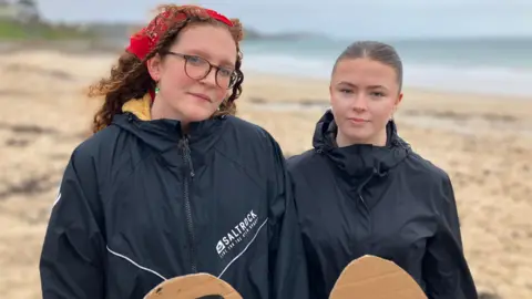 Jasmine Boniface and Verity Jones are looking serious as they stand on sandy Gyllyngvase Beach with the sea behind them. They are both wearing black anoraks and are holding cardboard placards but we cannot see what is written on them. Ms Boniface has dark red corkscrew curls and is wearing a bandanna in her hair and a pair of glasses. Ms Jones has her light brown hair pulled tightly back off her face and has multi-studded ears and a nose ring.