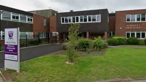 A two-storey school building with a mix of red brick and tiled frontages. A sign reading Aldersley High School in cut-out metal lettering is on the wall to the right, while a white sign stands on the grass in front of the building reads Welcome to Aldersley High School, directing visitors to reception. 