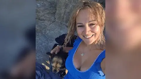 Handout Kayleigh is wearing a blue swimsuit and sat by some rocks on a coastline with a small black and brown dog beside her. She has blonde hair and is smiling at the camera.