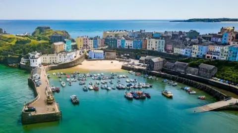 Getty Images About 80 small boats moored in green-blue water in the harbour in Tenby, viewed from above in bright sunshine. There are rows of multi-coloured houses behind the harbour - green, blue, yellow, turquoise, orange and pink - and a view out to sea at the top of the frame.