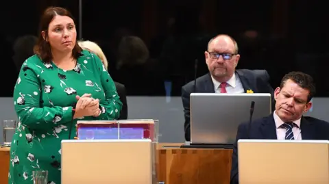 Welsh government Heledd Fychan has short brown hair and a green dress with black and white flowers on. She is sat next to Rhun ap Iorwerth in the Senedd siambr