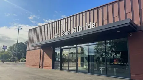 Network Rail Exterior view of East Kilbride railway station building with ScotRail branding, featuring modern architecture with large glass doors and a brown metal facade under a partly cloudy sky.