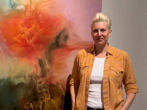 BBC/Emma Richardson A smiling Emma Richardson is wearing a camel coloured overshirt with a white t-shirt underneath. She's standing in front of her own oil painting which merges red, orange, green paint colours in a striking, largely blurred pattern. 