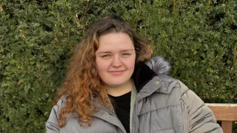 George King/BBC A young woman with long curly hair wearing a grey jacket. She is sitting on a bench and smiling into the camera