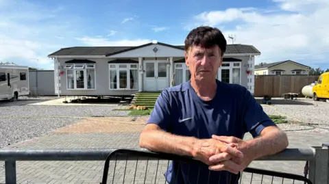 Ben Schofield/BBC Clifford Codona leaning on a gate and looking direct to camera. He is standing outside, with a static caravan in the background. While the caravan is centred, Codona is positioned just to the right of centre. His forearms are resting on the top of the gate and his hands are clasped together in front of him. He is wearing a blue T-shirt. He has dark hair with greying temples.