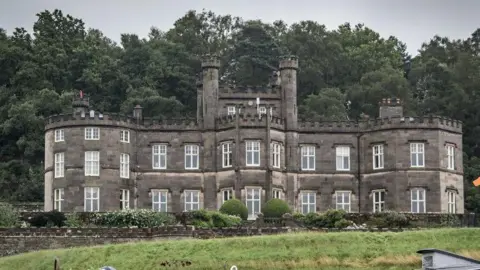 A large grey building with a crenellated roof line, a turreted central tower and white sash windows.