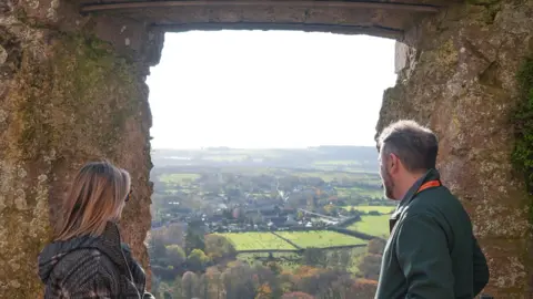 A woman on the left and a man on the right both peering out of a lookout from the old castle walls. Fields, housing and trees can be seen in the distance as they look out on the scene.