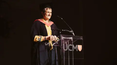 MATT MOSUR Gail Corrin a woman with short hair, wears a graduation robe and hat as she makes a speech in front of a Perspex podium with UCM on the front. 
