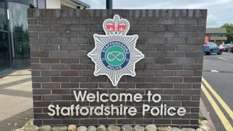 A brick wall outside Staffordshire Police headquarters. The wall has the force's badge on it, a silver star shamed symbol with a red crown and a silver rope on a green background, and the wording 'Welcome to Staffordshire Police'