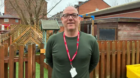 Andy Robinson, who is bald, wears black framed glasses and a green long-sleeved top. He is wearing a red lanyard around his neck and is smiling at the camera in front of a wooden fence enclosing a play area.