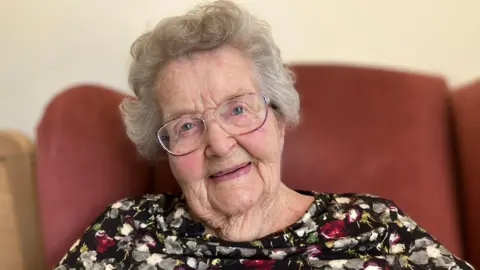 Ula Rigg sitting in a red armchair. She has round glasses on and grey hair, and a floral top
