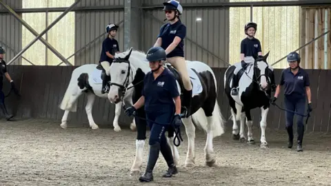 A group of three young people are led on black and white piebald ponies around an indoor riding arena by instructors.