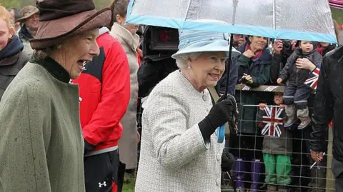Getty Images Queen Elizabeth II, dressed in a cream coat and hat and holding an umbrella, is accompanied by Dame Shân Legge-Bourke, in a green coat and brown hat. There are cheering crowds with flags in the background. 