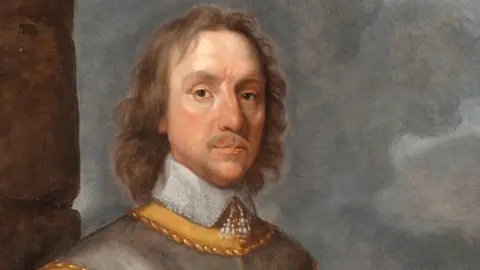 A 17th Century portrait of Oliver Cromwell, with shoulder-length curling mid-brown hair and moustache, wearing a white lace collar done up at the neck over a yellow under-jacket, above which is grey armour. He looks at the viewer with dark eyes.