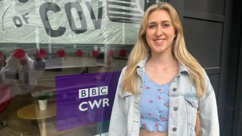 A blonde woman wearing a blue cropped shirt and a denim jacket. She is smiling at the camera 
