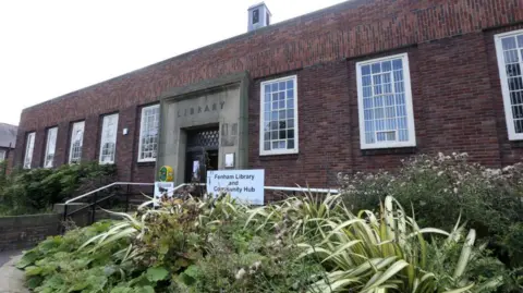 Fenham Library and Community hub. The single-storey building is made from red bricks and has a lot of plants growing outside. Steps lead to the building's entrance which is under a large stone arch in the building.