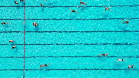 An aerial view of a bright blue swimming pool which has long black lines running along the bottom. People are swimming in the pool, some stand