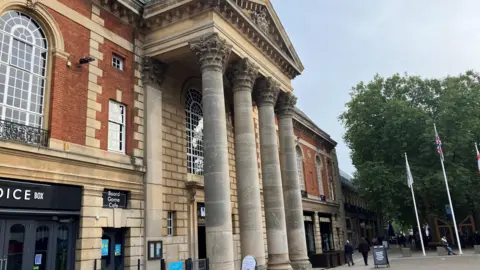 The exterior of Peterborough Town Hall, with tall stone columns providing a grand arch over the entranceway. There are shops, shoppers and flagpoles also in view.