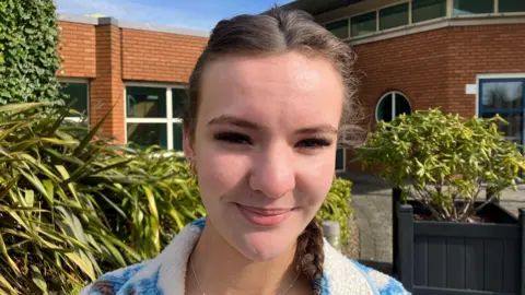 Georgia is standing outside. A school building and foliage is behind her.Her brown hair is in a plait. She's wearing a fleecy jacket in blue and white. 