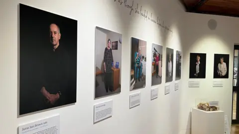 A number of rectangular portraits of staff and volunteers of Les Bourgs Hospice displayed on a plain white wall. They each have an informational plaque below each one. 