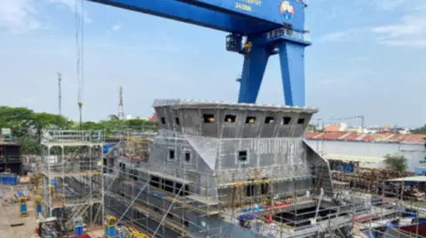 Scilly Steamship Group A huge crane is seen over the skeleton of a grey vessel with scaffolding in the foreground and houses in the background