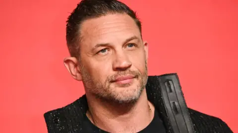 Getty Images Tom Hardy wears a black cardigan. He is stood in front of a red background