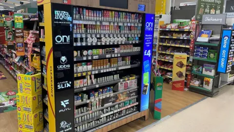 A large display of vapes at the end of a Halloween sweets aisle in Morrisons in Worle. It is a large display with colourful vape boxes and electronic screens.