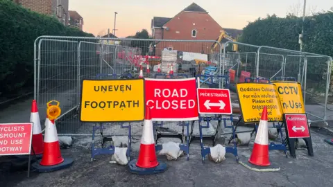Mark Carter/BBC Wire fences surround a roadworks side. Multiple red and yellow warning signs and some traffic cones have been set up in front of the fences.