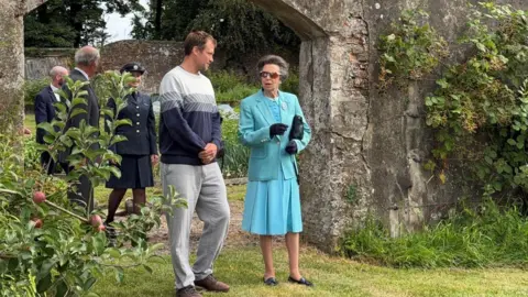 Princess Anne wearing a teal outfit. She is standing next to a man with short brown hair wearing a grey jumper. Princess Anne is wearing sunglasses and gloves. Behind them are three people. Princess Anne and the man next to her are standing on a patch of grass in front of a stone archway. There are ushes around them.