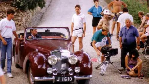 Kevin Lewis A colour photo from the 1980s. People on the Bergerac TV set are stood, and sat, on a road in Jersey, crowded around John Nettles driving a burgundy car. Kevin Lewis is stood to the left of the car with his hand on the side of it.