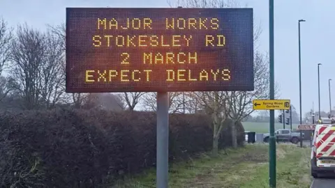 An illuminated sign at the side of the road warning of delays due to roadworks at Stokesley Road.