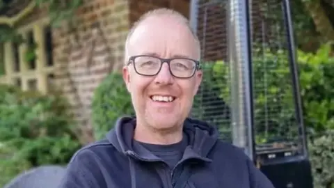 Family photo Damian Barr smiles in an outdoor cafe with shrubs behind covering a wall. He wears dark glasses and dark hooded top. 