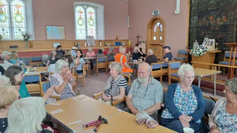 Sandra Pratt Inside the church building. Tables and chairs have been placed around the round the room. A group of people are scattered around the room, socialising. They are sitting on chairs and are chatting. They appear to have raffle strips on the table in front of them. The room has pink walls and large stained-glass windows.