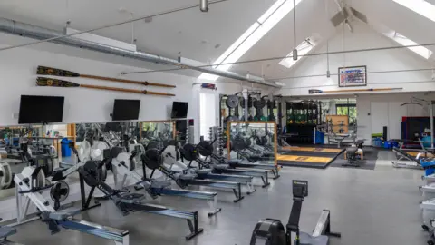 David Johnson Photographic Rowing machines are lined up inside a boathouse and there is other gym equipment in the large room.
