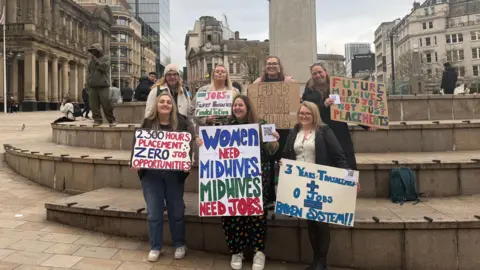 Seven student midwives stand together holding signs protesting the lack of jobs. The are standing on steps in a square. The placards they are holding read "Women need midwives, midwives need jobs", "2,300 hours placement, zero job opportunities", "Three years training and 0 jobs = broken system", "Future midwives need jobs not placements".