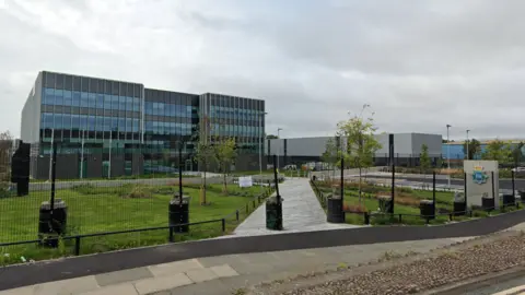 Google Merseyside Police HQ, a large rectangular glass fronted building with grey cladding, and a grassy area and car-park behind a fence in the foreground.