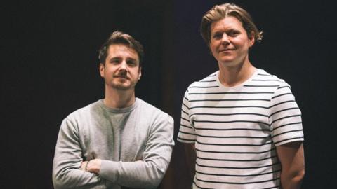 Two men, one shorter man in a grey jumper with brown hair, and a taller man with a stripy t-shirt and blond hair.