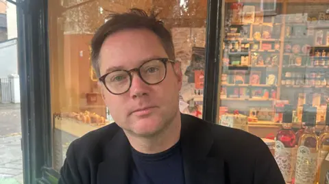 A man with short brown hair and round dark glasses looks at the camera