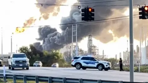 A large grey plume of smoke rises from an oil refinery.