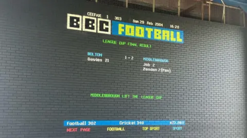 Mural in the style of the former television teletext service Ceefax. Set against a dark grey brick wall it states in red, yellow, green, blue and white letters: BBC Football: League Cup Final Result. Bolton 1 -2 Middlesbrough. Middlesbrough lift the league cup
