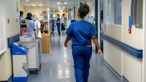 PA Media A nurse in blue overalls walks along a hospital corridor with her back to the camera . A number of other medical staff can be seen in the background.