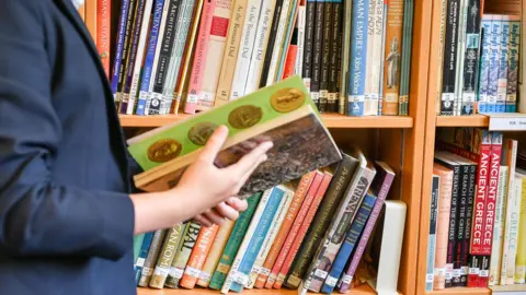 A person wearing a navy blazer holding a book in her hand. Behind are rows of books in a library.