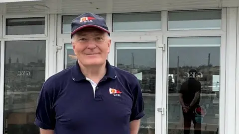 A man in blue cap and blue polo shirt smiles at camera standing in front of Guernsey Yacht Club.