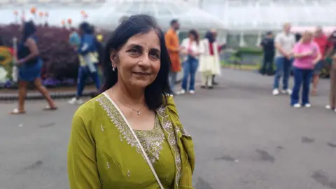 Dr Nisha Tandon is dressed in green. She has black hair. She's standing in the Botanic Gardens with a hothouse behind her. People are behind her in the background.