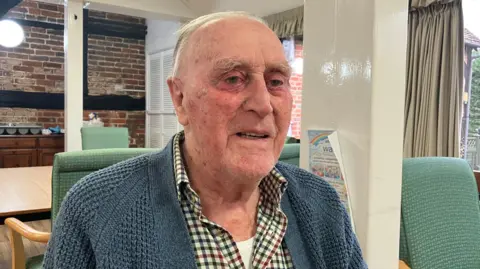 William has short white hair. Like his fellow day centre visitors he is sat on a chair in a lounge area.