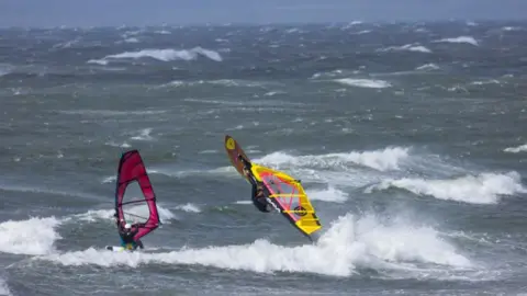 Jonathan Head Two windsurfers in waves, one with a yellow sail is airborne, with a rough sea behind them.