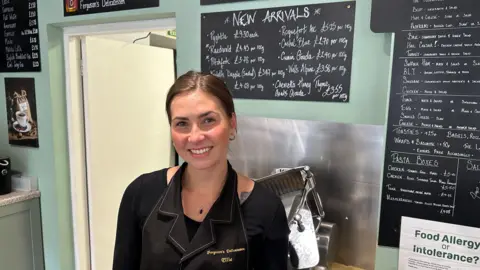 Ben Parker/BBC A woman wearing a black top and black apron, she is standing in front of chalkboards with items and prices written on.